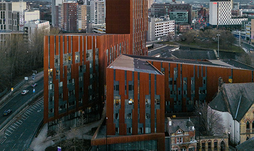 Aerial view of the University of Manchester’s red-toned engineering building at sunset, with surrounding roads, modern high-rises, construction cranes, and city skyline in the background.