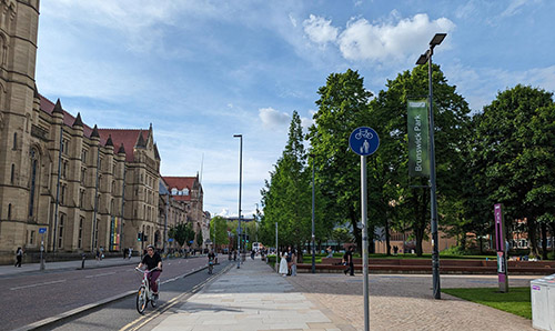 Street view of Oxford Road in Manchester showing cyclists, pedestrians, the Whitworth building with red roofs on the left, and Brunswick Park with green signage and trees on the right.