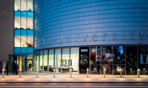 Evening view of The Place building at The University of Manchester, with illuminated interior spaces and science themed posters displayed on the ground floor windows facing the street.