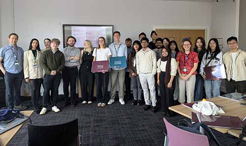 Group of students and faculty posing in a classroom setting, with some holding project folders labelled WOODLEY and DANE ROAD, under the Manchester Urban Design Lab as seen on the screen behind.