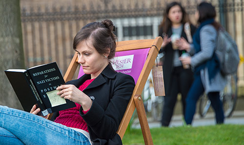 Student reading a textbook on a chair outside.